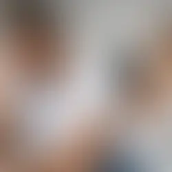 Hombre preocupado sentado en cama, con mano en la cabeza. Al fondo, mujer sentada de espaldas. Flores en la cama. Ambiente tenso.