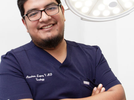 Hombre con gafas y barba sonríe, lleva uniforme azul médico. Está bajo una lámpara quirúrgica en un ambiente clínico.