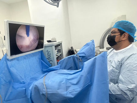 Persona con mascarilla observa una pantalla médica en un quirófano. Ambiente aséptico con equipo azul. Monitoreo de procedimiento.