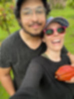 Two people smiling in a green garden. One holds a red cocoa pod, wearing red sunglasses and a colorful cap. Both wear black shirts.