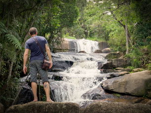Homem de costas observa uma cachoeira em degraus na floresta, com tripé ao lado e clima de aventura.