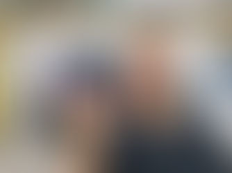 Homem surpreso em home office, lançando um dispositivo calibrador de monitor preto; monitor ao fundo, prateleira de livros e quadro de onda.