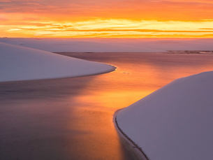 Dunas de neve brancas ao pôr do sol, com água refletindo tons laranja e céu dourado, cenário calmo e amplo.