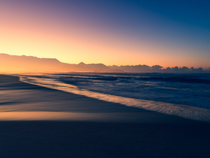 Mar ao amanhecer com ondas suaves, montanhas ao fundo e céu em degradê laranja e azul, clima sereno.