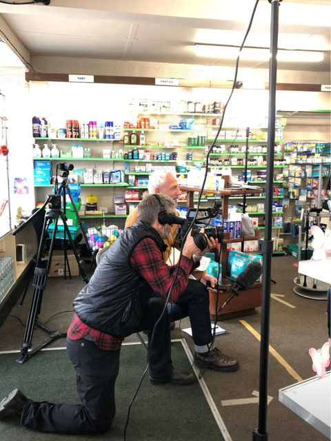 Photographer filming a man with a camera in a pharmacy aisle setting.