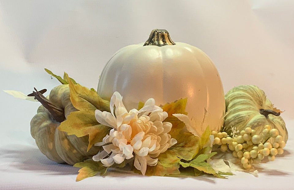 A small off-white faux pumpkin with smaller green pumpkins, leaves and white mums at its base.