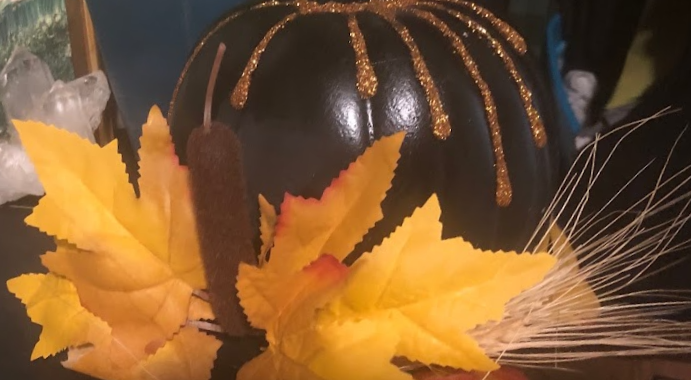 A small faux black pumpkin with gold glitter drips running from the crown to a base of yellow leaves and faux cat tails.