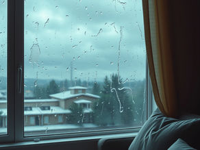 Raindrops on a window with a view of cloudy skies and houses. A cozy sofa with blue cushions and yellow curtains adds a calm mood.