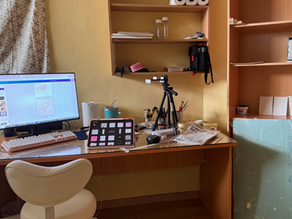 Neat craft desk with monitor, pink keyboard, tripod, shelves of supplies, and a white chair in a warm yellow room.