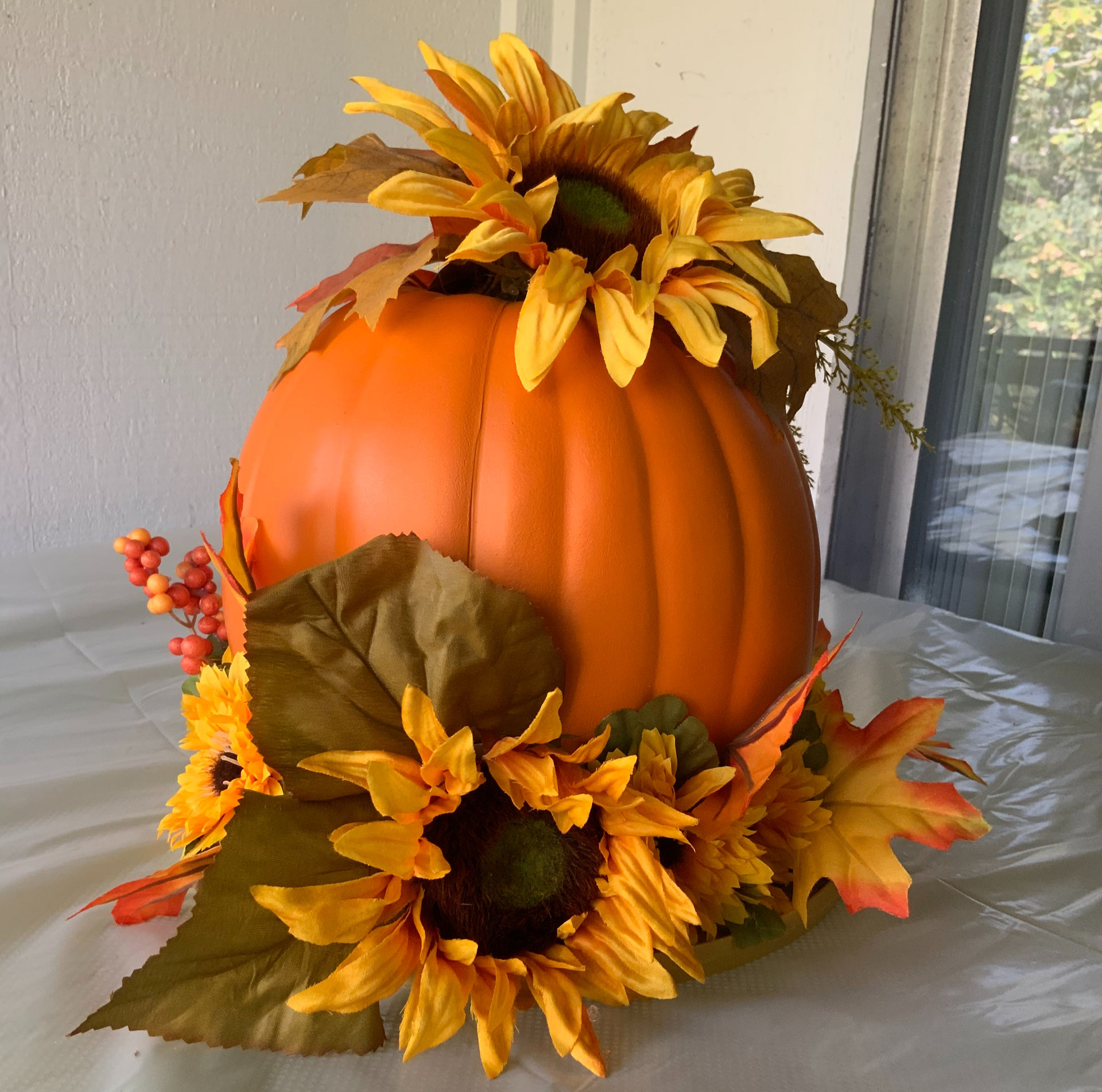 A faux orange pumpkin sitting in a base of sunflowers with a sunflower crown.