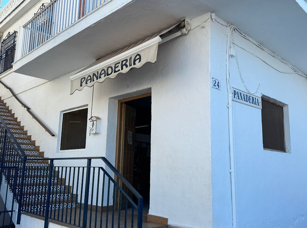 White building with "Panadería" sign, open door, and blue-accented staircase. Address number 24. Sunny day, inviting mood.