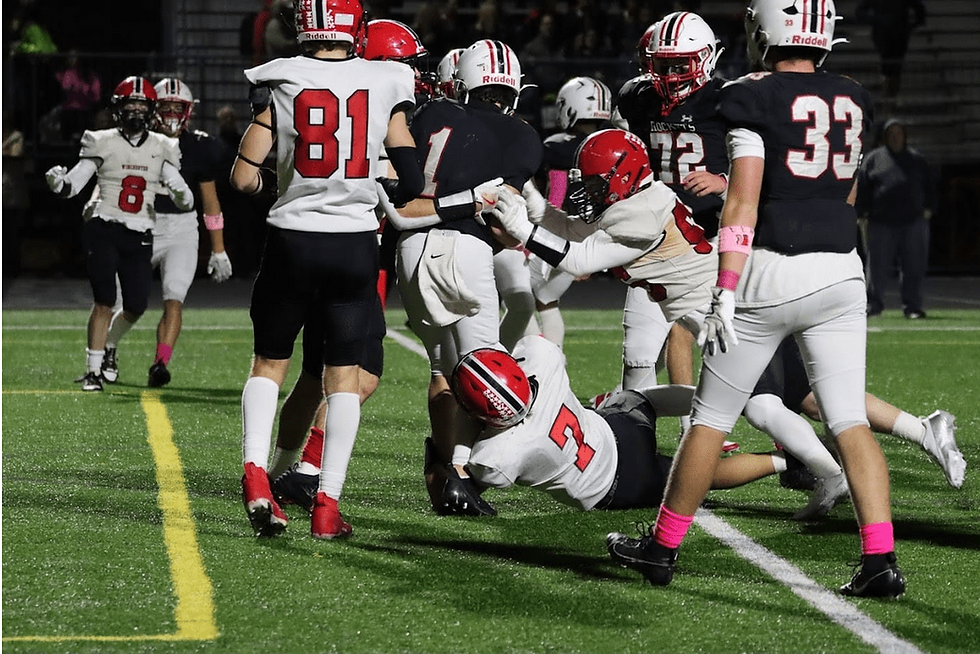 Football game action, players in red and white uniforms; one tackles another on a green field. Intense focus, crowd blurred in the background.