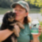 Woman holding a bottle of NuVet Plus canine wafers while cuddling a friendly tricolor Poodle puppy outdoors by a lake, highlighting daily dog nutritional support and immune health supplements.