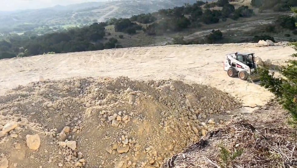 Bulldozer on dusty hillside, moving dirt. Mountainous landscape in background with green trees and clear sky, conveying a rugged, natural setting.
