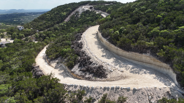 Winding dirt road curves through lush green hills under a clear sky. The landscape is serene and expansive, with distant views.