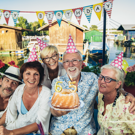 Group of five people in party hats celebrate outdoors by a lake. One holds a cake with "65" candles. Happy Birthday banner above.