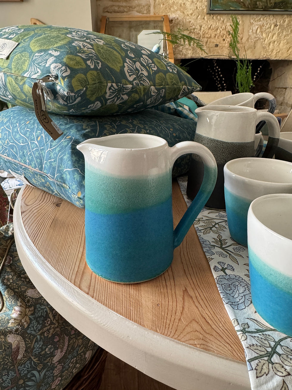 Jug with blue green and cream colours on wooden top with cushions behind