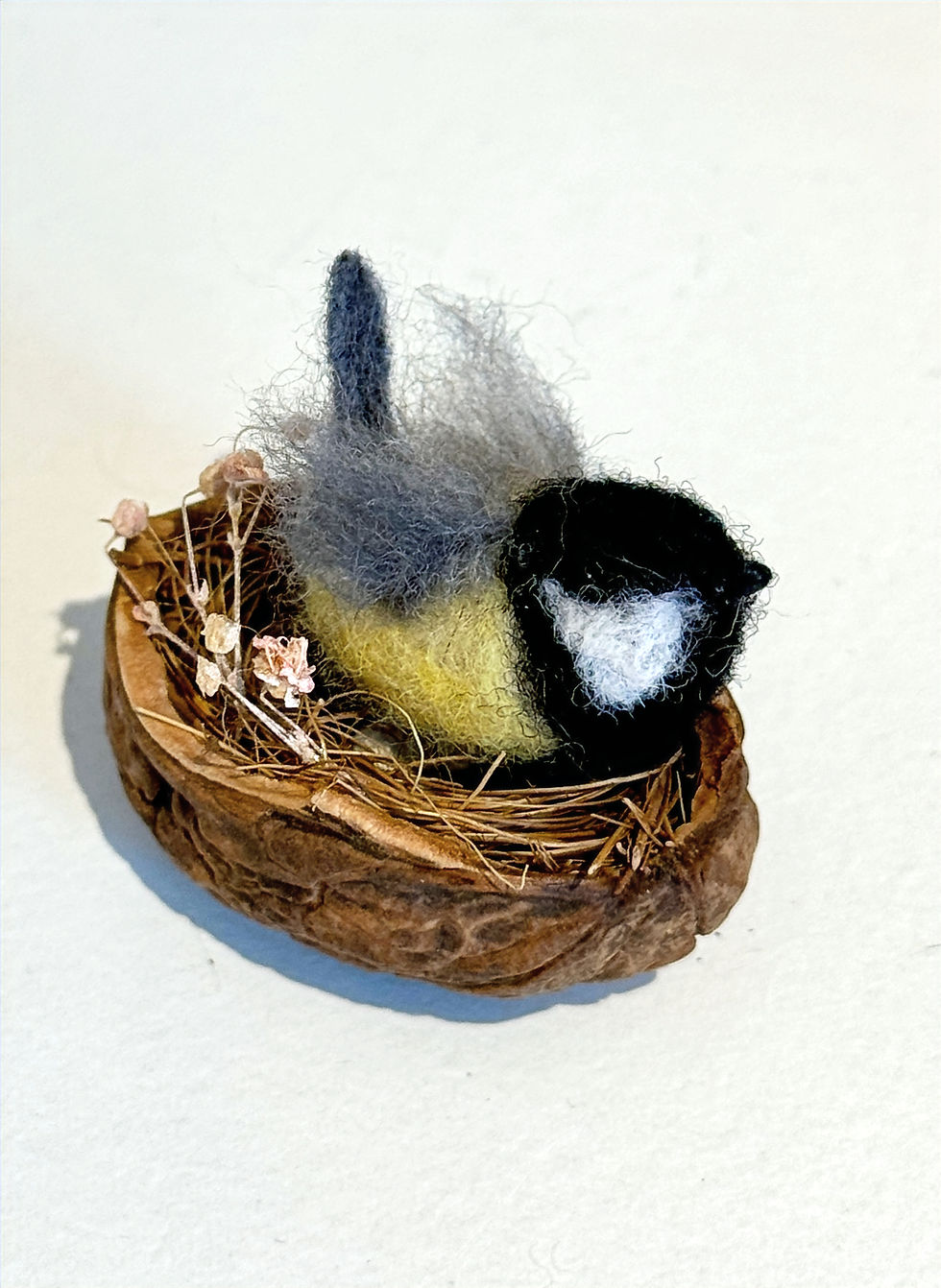 felted great tit with yellow black and grey sitting in a nest on a white shelf
