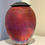 Thumbnail: Large jar in strong colours of red, orange, purple with black lid on white shelf