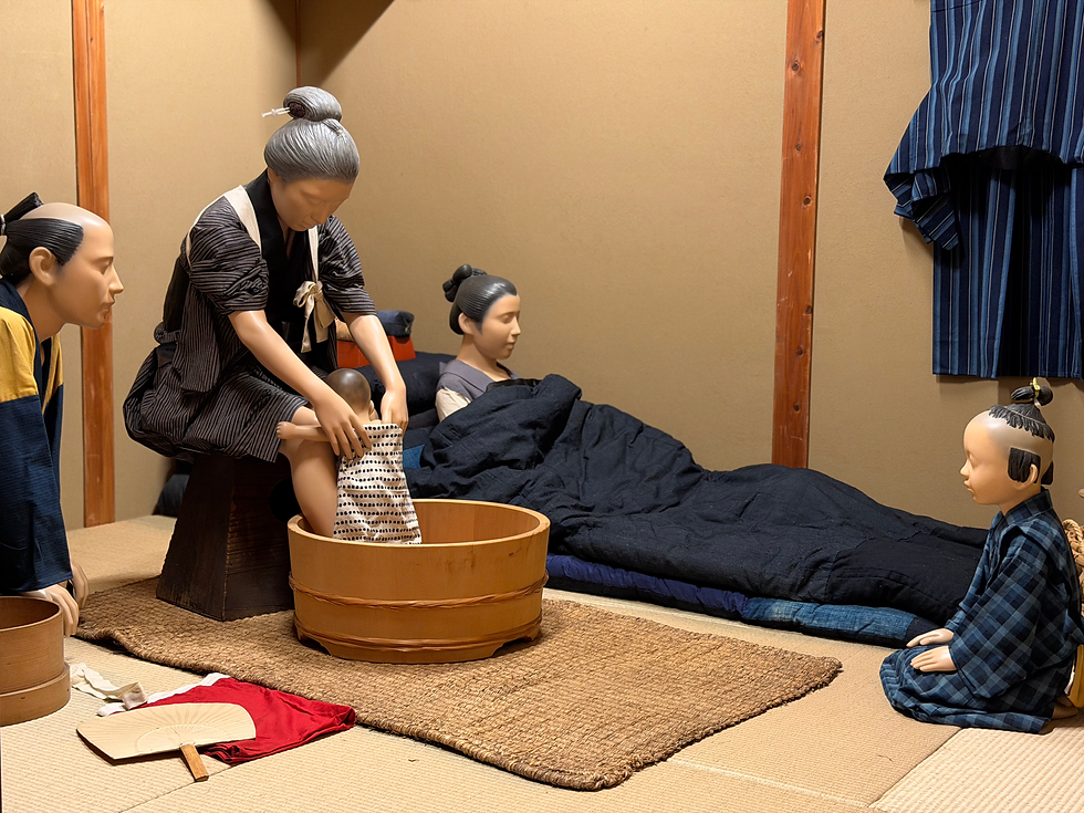 Bonecos em um quarto japonês, uma mulher lava um bebê em uma bacia de madeira, dois observam, roupa azul pendurada na parede.