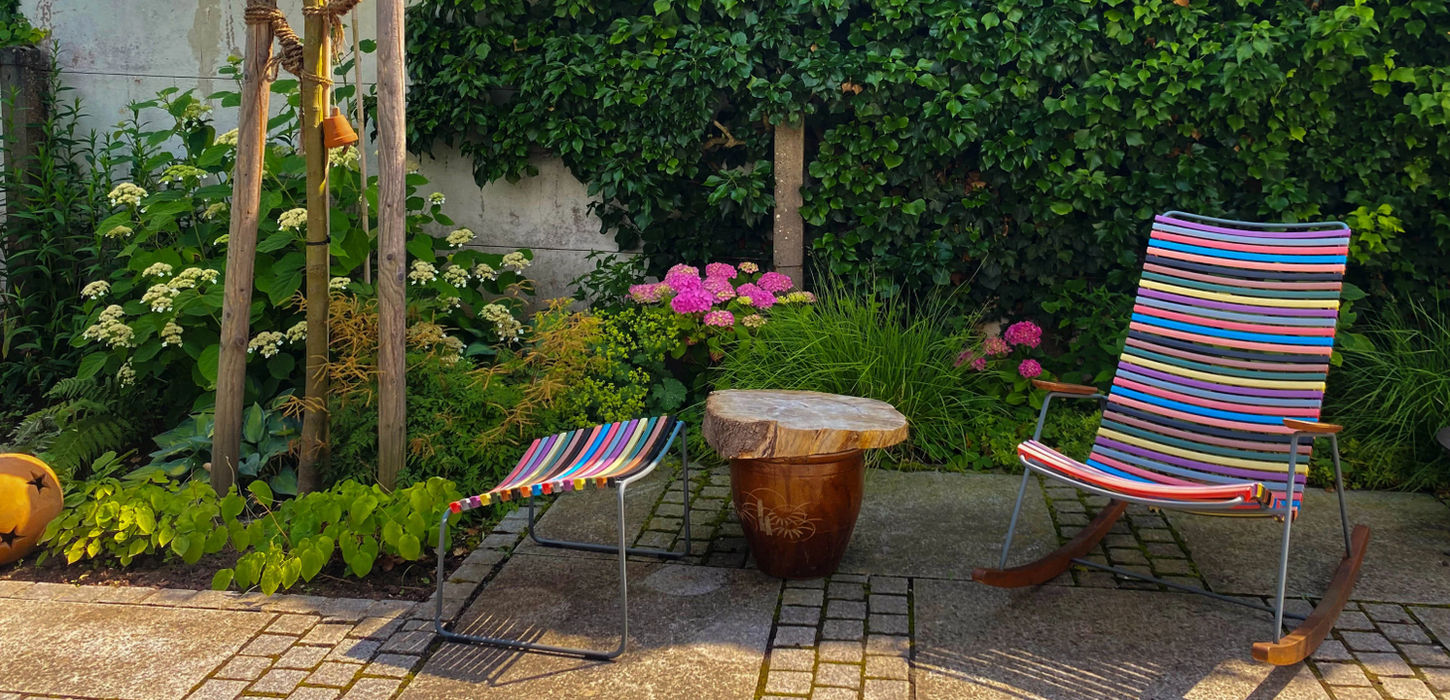 Gemütlicher Sitzplatz mit in allen Farben bespanntem modernem Schaukelstuhl und Hocker umgeben von weiß blühenden Hortensien, Efeu und Blattschmuckstauden