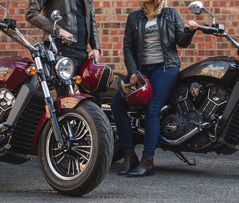Studio photography of a man and woman in leather jackets posing with cruiser motorcycles. The scene has an adventurous, edgy, and stylish mood against a brick wall background.