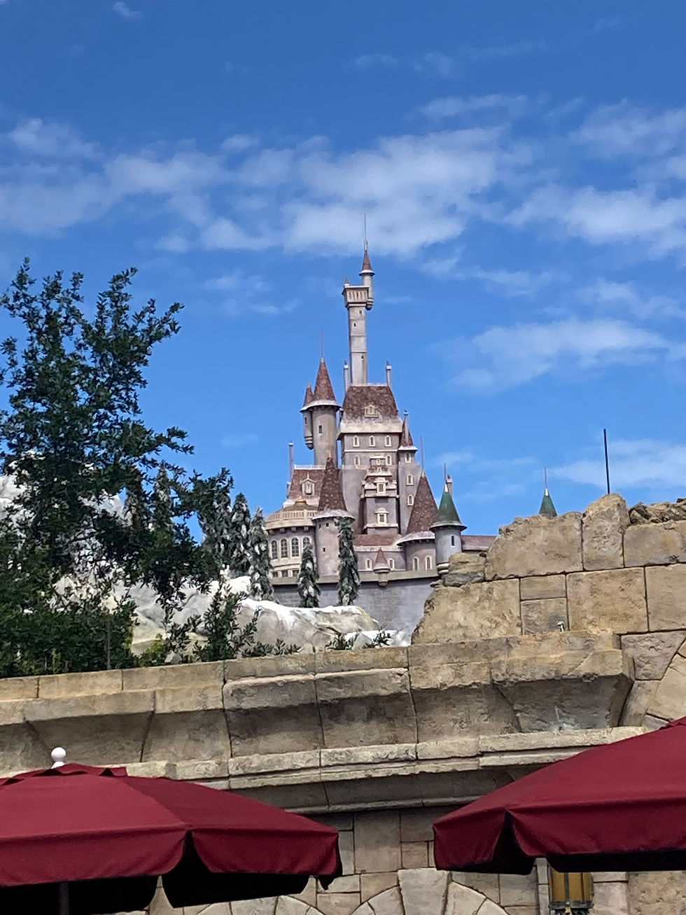 A fairytale castle with towers rises against a blue sky. Foreground has stone walls and maroon umbrellas, creating a whimsical scene.