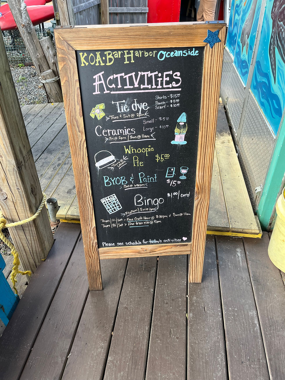 Chalkboard sign for KOA Bar Harbor Oceanside activities lists tie dye, ceramics, whoopie pie, BYOB paint, and bingo on a wooden deck.