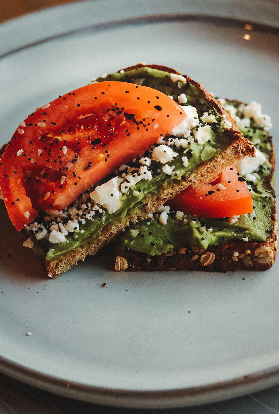 Avocado Toast on Dave’s Killer Bread topped with feta cheese and freshly sliced tomatoes, served on a rustic wooden table at Harvest Coffee.