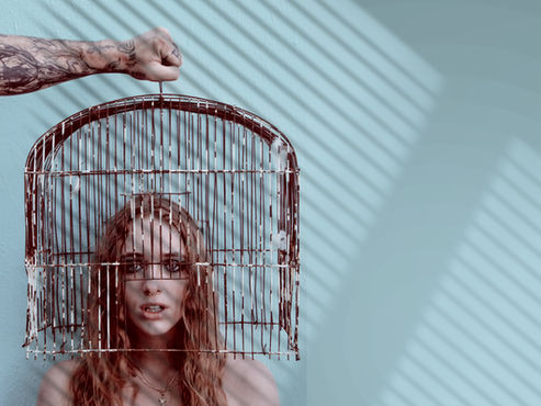 A tattooed arm holds a birdcage over a woman's head against a teal wall with striped shadows. She looks pensively at the camera.