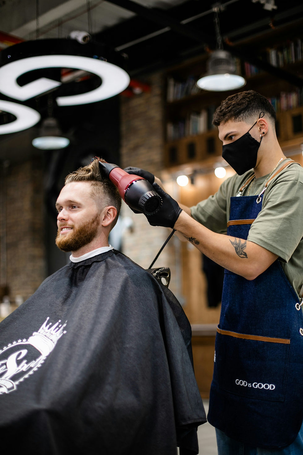 Barber in mask uses hairdryer on smiling client. Black cape, visible tattoo, "GOD IS GOOD" apron. Modern, well-lit barbershop.