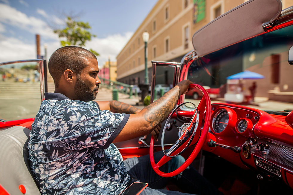 Man in a floral shirt drives a red classic car. Urban background with a sunny sky. Focused expression, vibrant colors.