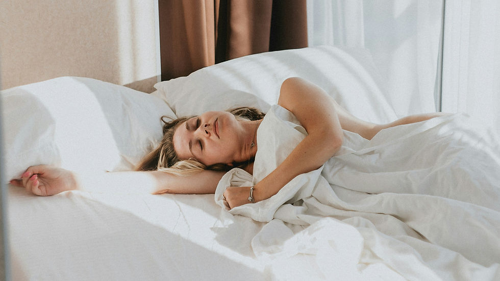 Woman sleeping peacefully on a sunlit bed with white sheets. Brown curtains in the background create a warm, relaxed atmosphere.