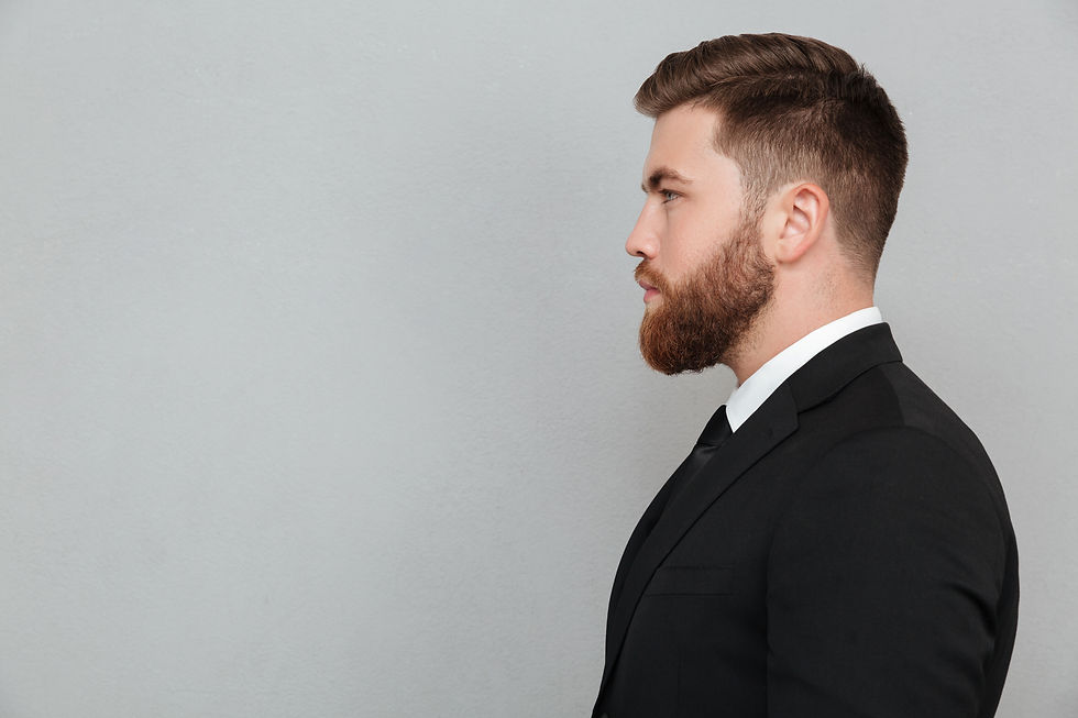Man in profile wearing a black suit and white shirt, with a groomed beard, standing against a plain gray background. Neutral expression.