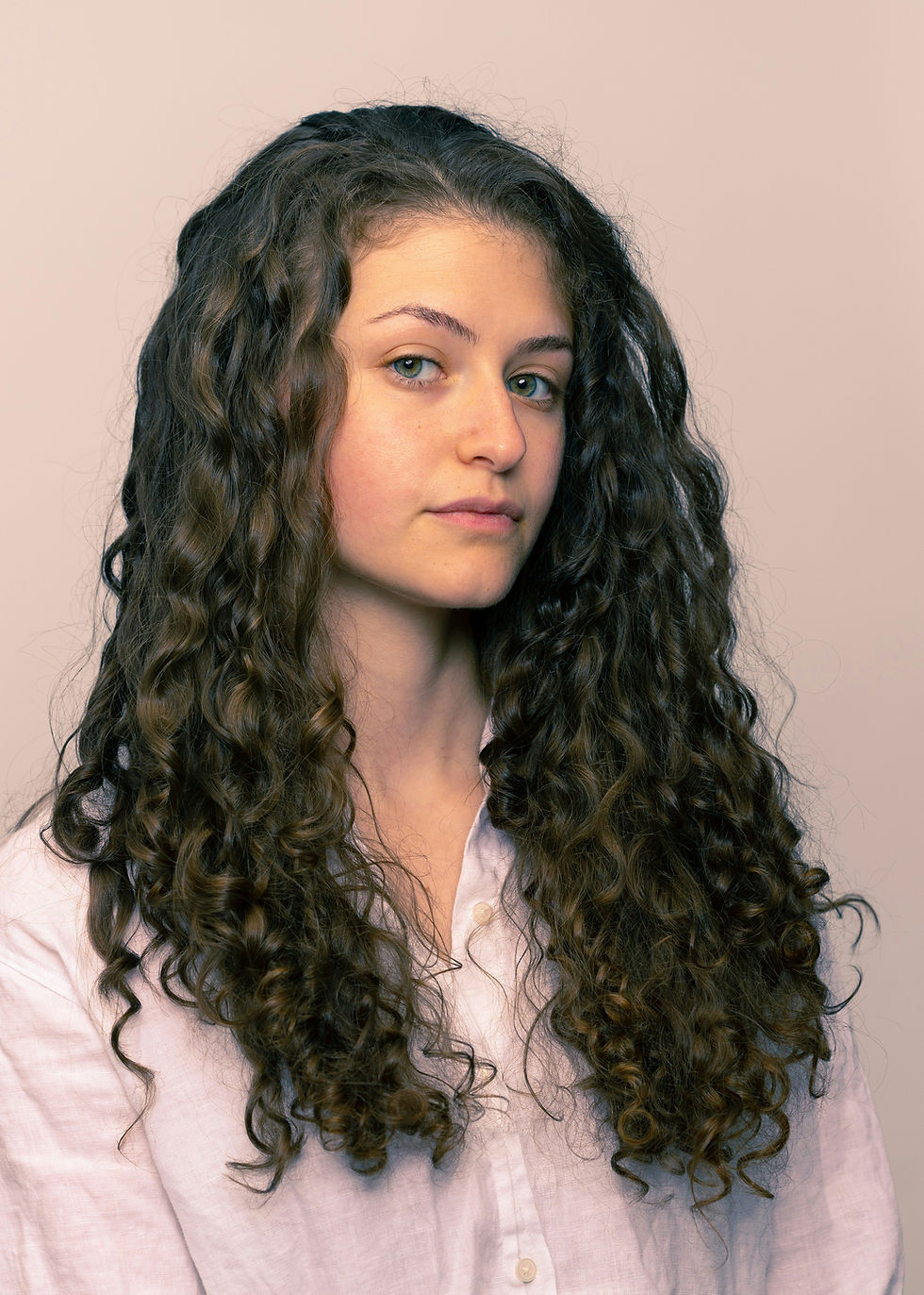 Young person with long curly hair and a neutral expression wearing a white shirt against a soft beige background.