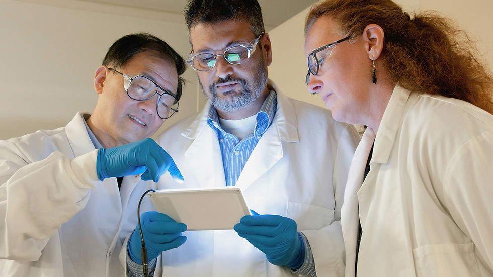 Three scientists in white lab coats and blue gloves examine a tablet together, focused and engaged in a brightly lit lab setting.