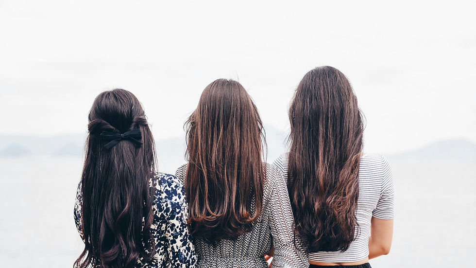 Three women with long hair stand by the sea, wearing patterned dresses. Overcast sky and calm water set a serene mood.