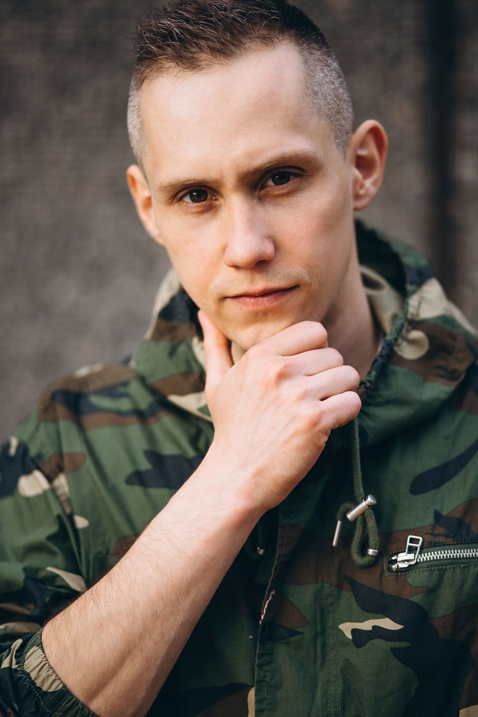 Man in a camo jacket thoughtfully touching his chin against a blurred background, conveying a reflective mood.
