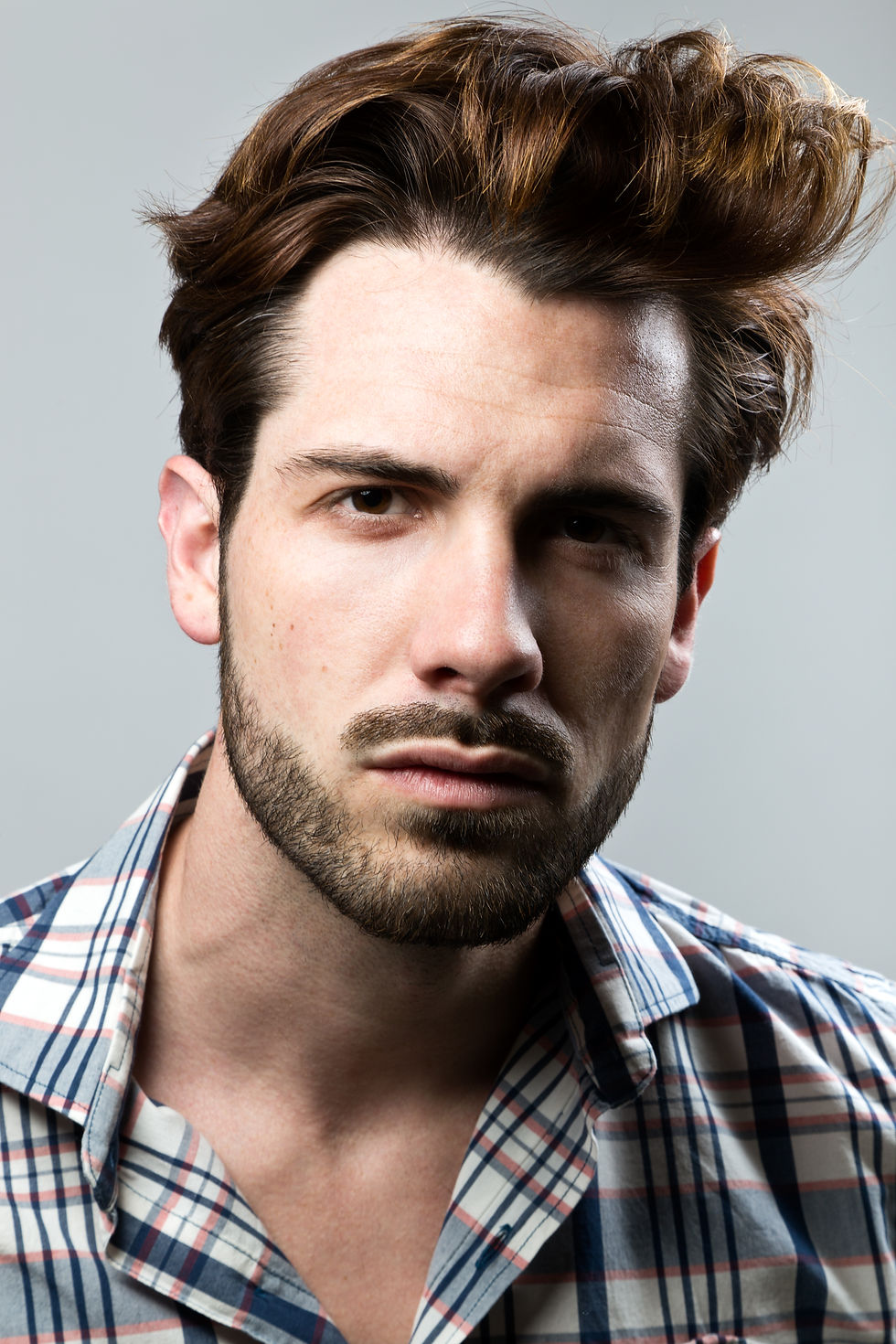 Man with styled hair and a beard in a checkered shirt gazes intensely at the camera. Gray background, neutral mood.