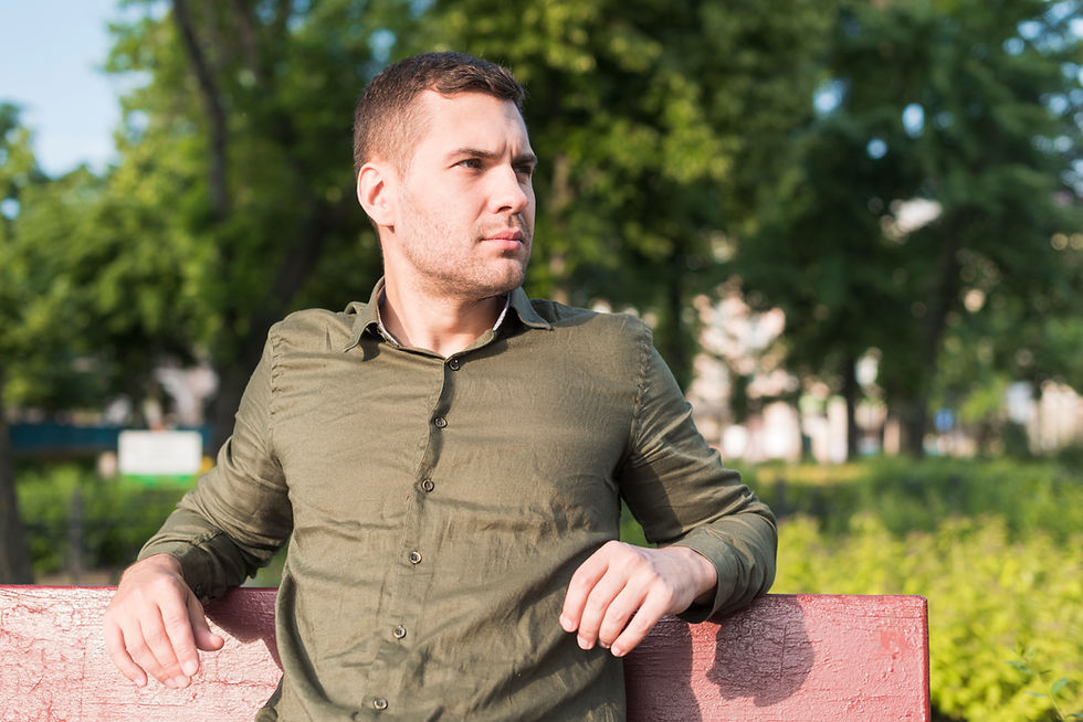 Man in a green shirt sits on a red bench, looking thoughtfully to the side. Lush greenery and trees in the sunny background.