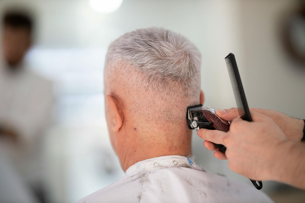 A person with short gray hair is getting a haircut with clippers in a bright barbershop. White cape, focused atmosphere.