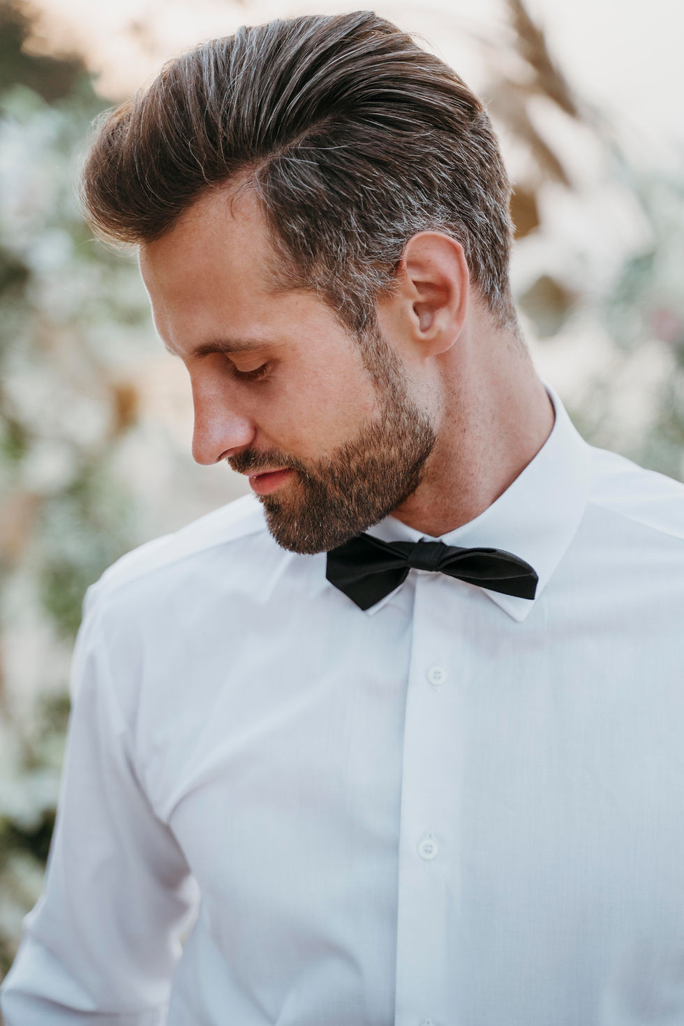 Man in white shirt and black bow tie gazes downwards with a serene expression, outdoors with a blurred natural background.