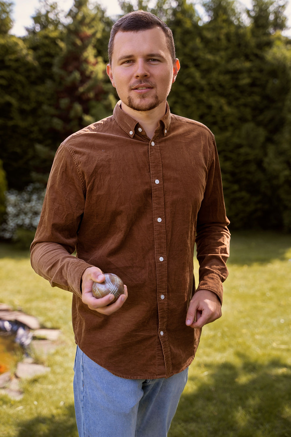 Man in a brown shirt holding a baseball stands on a grassy lawn, surrounded by lush trees, under a sunny sky.