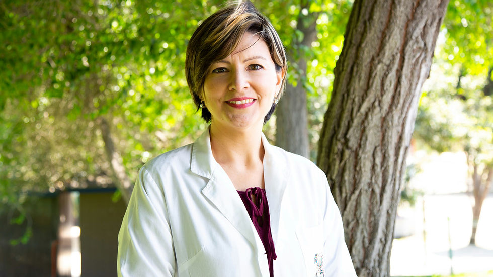 A smiling person in a white lab coat stands by a tree in a sunlit park. Lush green leaves form the vibrant backdrop.