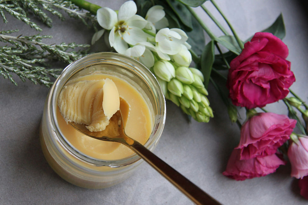 Jar of shea butter with a golden spoon, surrounded by white and pink flowers on a gray surface. Cozy and elegant vibe.