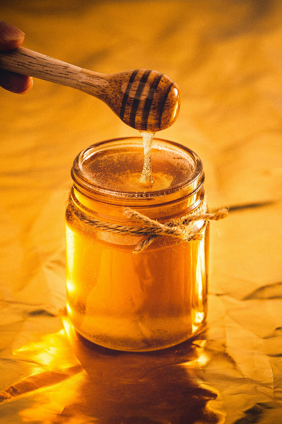 A jar of honey with a honey dipper, dripping golden honey. The jar is adorned with twine, set against a warm, amber-lit background.