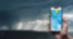 hand holding cellphone with moody clouds and ocean in background. on the phone screen is nagivation map.