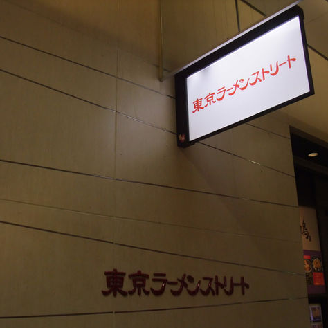 Illuminated sign on a beige tiled wall reads "東京ラーメンストリート" in red, indicating a ramen street location. Dimly lit interior visible.