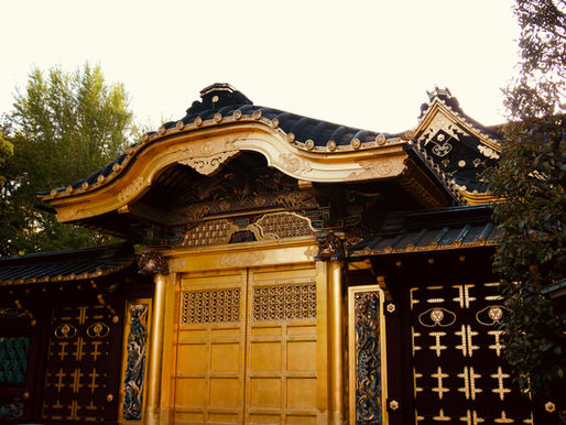 Ornate golden temple gate with intricate carvings against a clear sky. Dark roof tiles and decorative details, surrounded by green foliage.