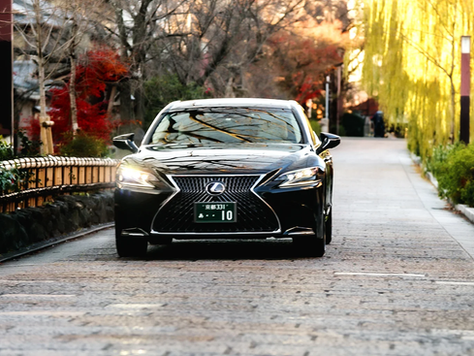 Black Lexus driving on a tree-lined street with autumn colors. Headlights are on; Japanese license plate visible. Road has a tranquil mood.